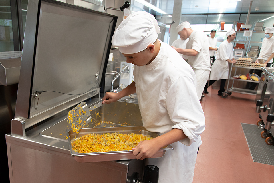 Das Bild zeigt einen Koch in der Grossküche beim Schöpfen von Gemüse.
