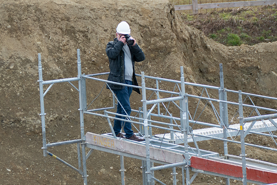 Das Bild zeigt einen Mann mit Bauhelm auf einem Baugerüst am Fotografieren.