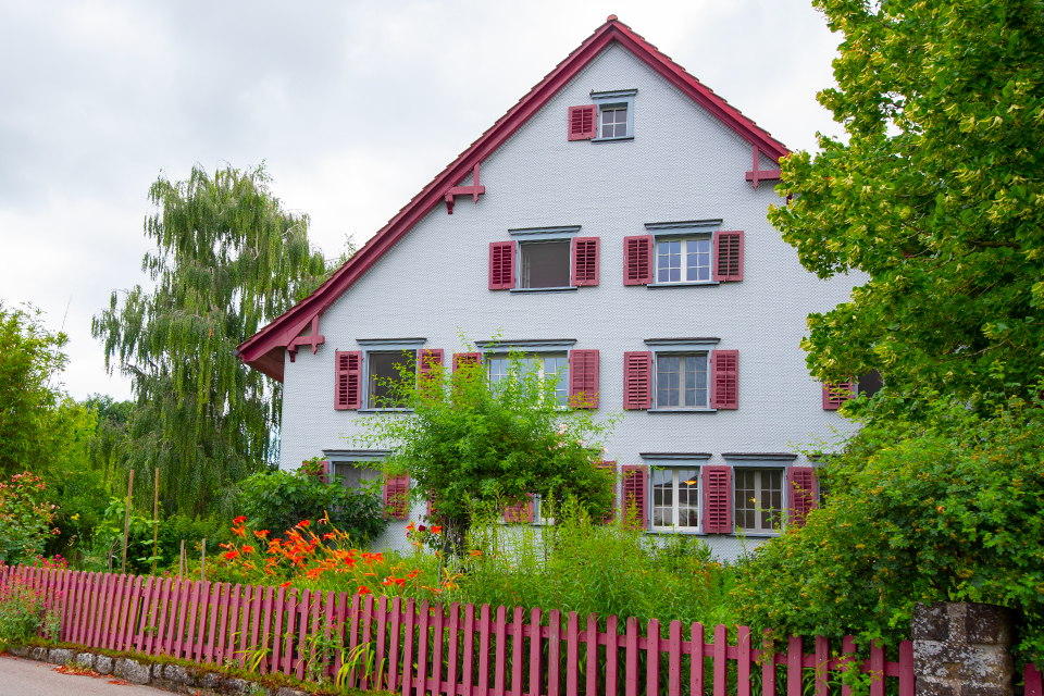 Das Bild zeigt ein grosses weisses Haus mit vielen Fenstern und roten Fensterläden. Vor dem Haus befindet sich ein grosser Garten mit Bäumen, Sträuchern und Blumen. Der Garten ist mit einem roten Gartenzaun eingezäunt.