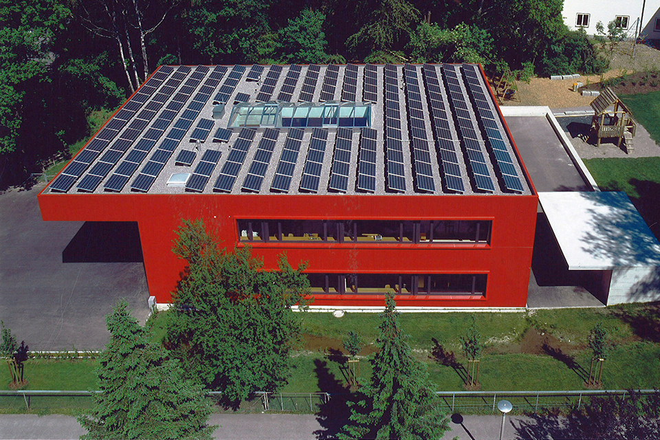 Das Bild zeigt die Vogelperspektive eines grossen roten Hauses mit Flachdach und Solarzellen auf dem Dach.