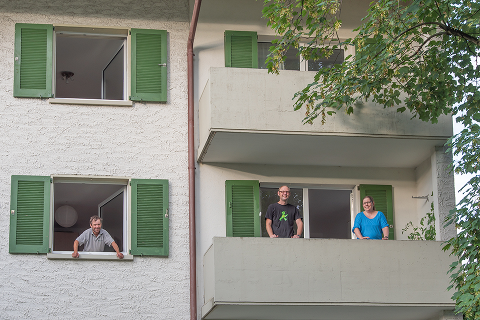 Das Bild zeigt einen Mann der aus dem Fenster eines Mehrfamilienhauses schaut und zwei Personen auf einem Balkon stehend.