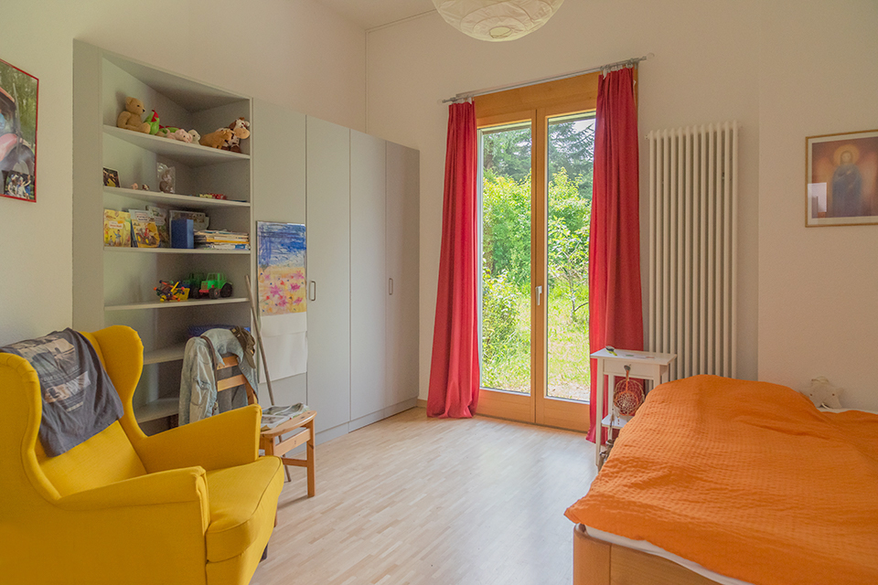 Das Bild zeigt ein Schlafzimmer mit Bett, Kleiderschrank und gelbem Sessel. Eine Glastüre führt in den Garten.