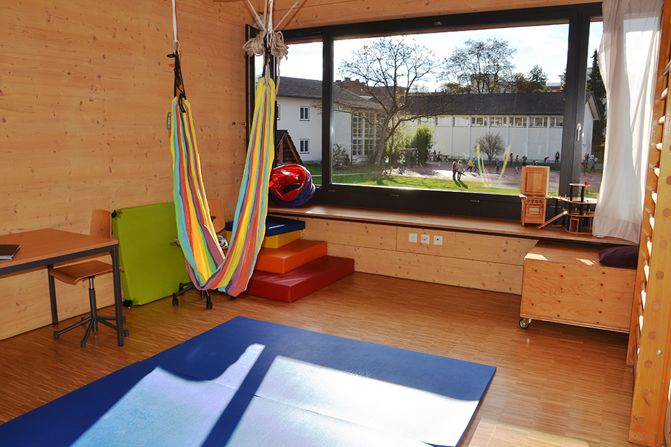 Das Bild zeigt ein Zimmer mit Holzboden und wänden und einem grossen Fenster mit Sicht auf den Schulhof. Im Zimmer befinden sich eine grosse blaue Turnmatte, eine Hängematte, eine Sprossenwand und ein Pult.