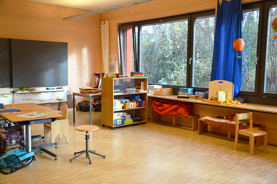 Das Bild zeigt einen Raum mit grossen Fenstern, einer Wandtafel und einem Pult sowie einem Regal voll mit Gesellschaftsspielen.