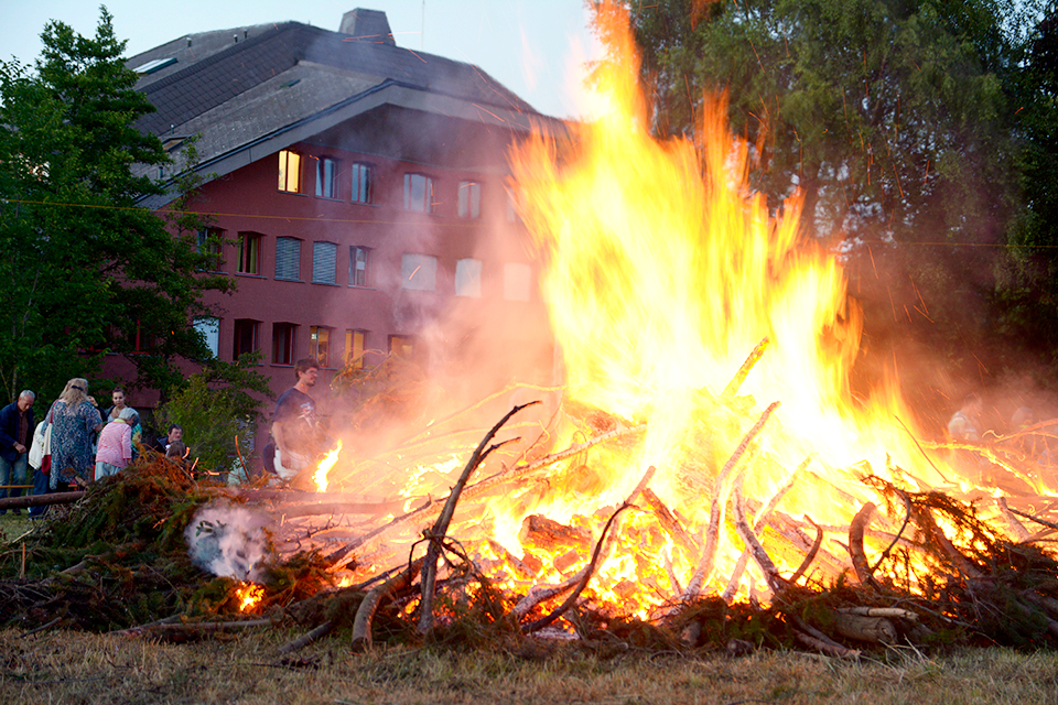Das Bild zeigt mehrere Menschen die um ein grosses Feuer stehen.