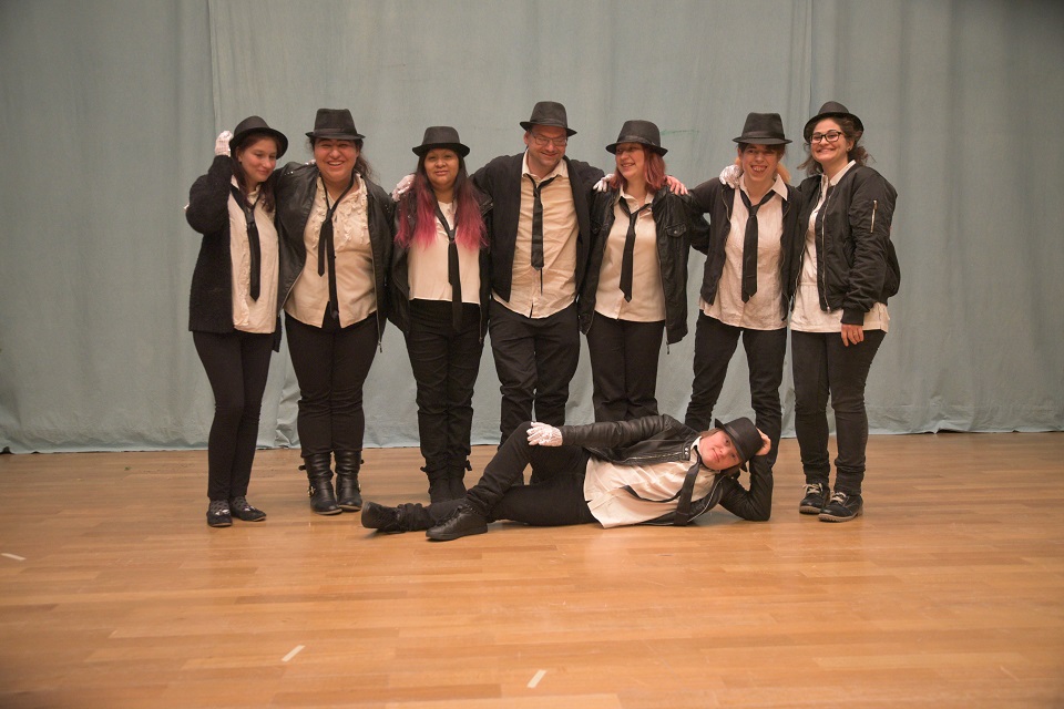 Das Bild zeigt acht schwarz weiss gekleidete Männer und Frauen die für ein Gruppenbild auf einer Bühne stehen.
