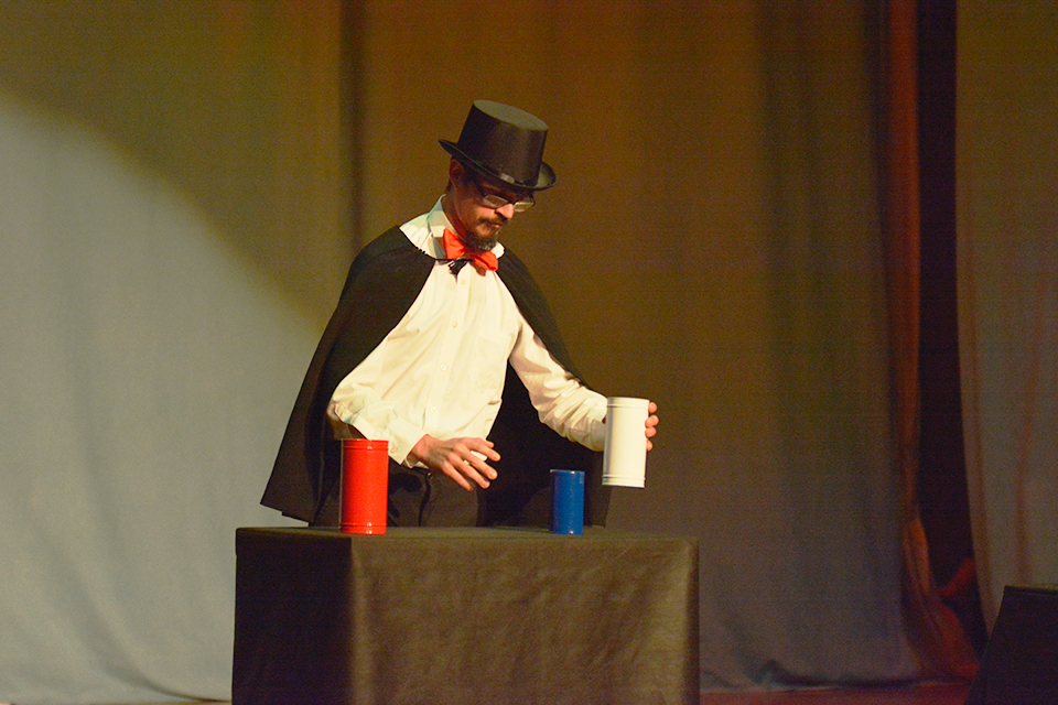 Das Bild zeigt einen Mann im Zauberkostüm beim Vorführen eines Zaubertricks mit drei Blechdosen.