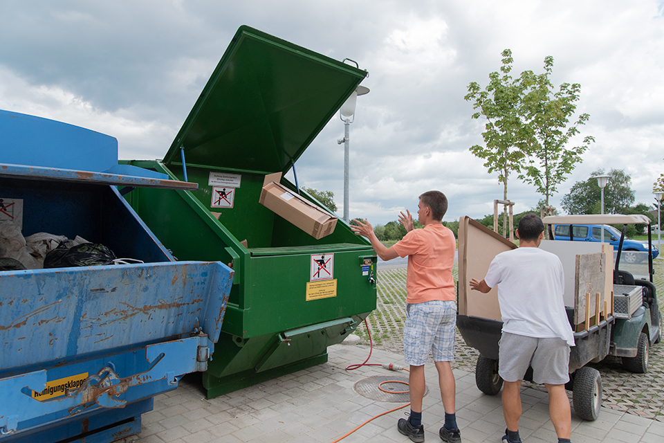 Das Bild zeigt zwei Männer beim Entsorgen von Kartonschachteln in Abfallmulden.