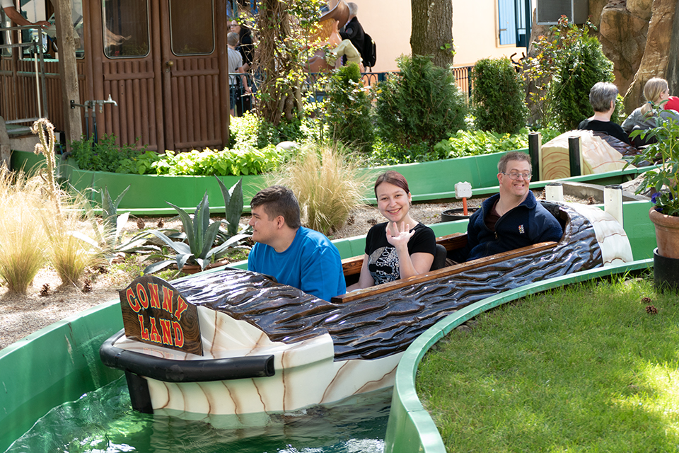 Das Bild zeigt eine Frau und zwei Männer in einem Freizeitpark. Sie sitzen zusammen in einem Boot im Wasserkanal.