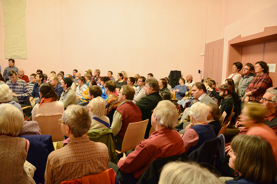 Das Bild zeigt einen grossen Raum mit vielen Menschen die auf einen Redner schauen.