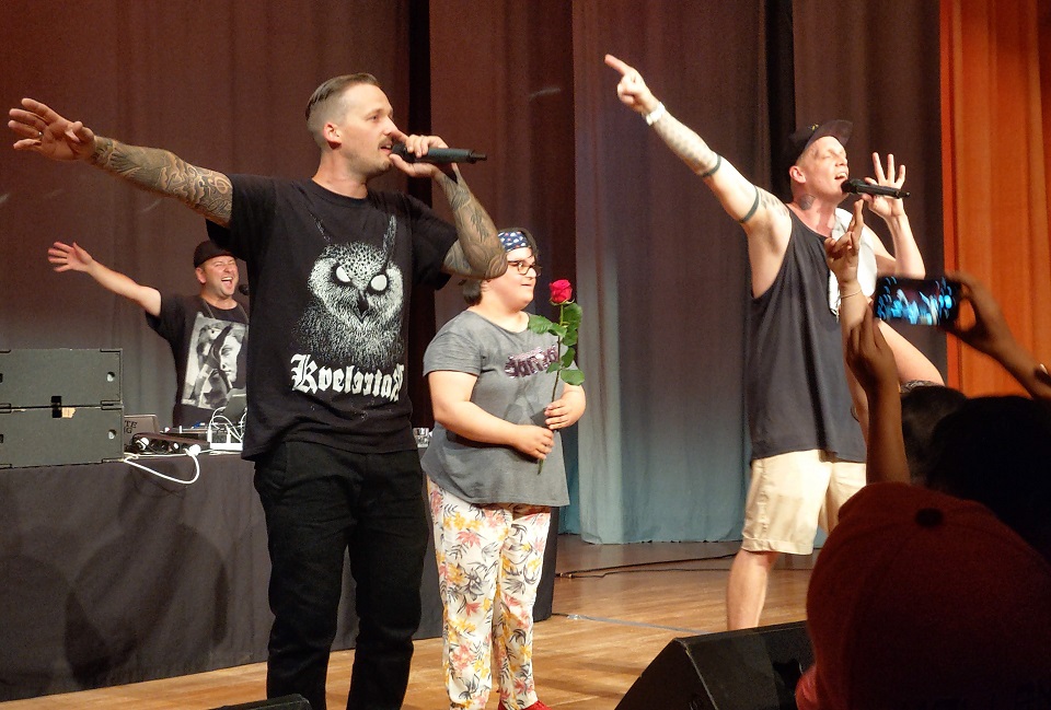 Das Bild zeigt einen DJ, zwei Rapper und einen junge Frau mit einer Rose in den Händen auf einer Bühne.