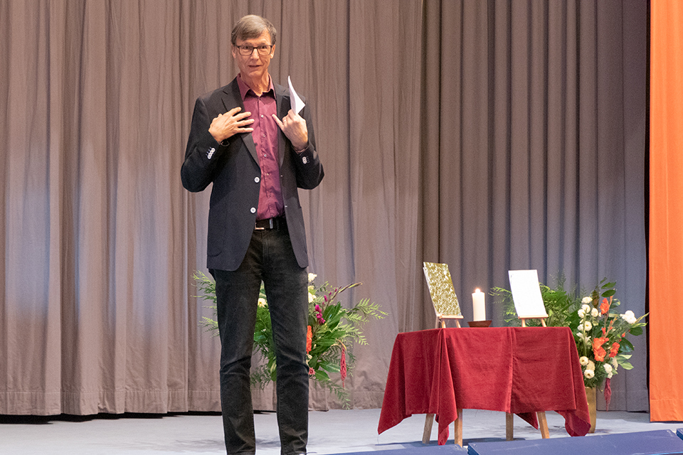 Das Bild zeigt einen Mann auf einer Bühne, der eine Rede hält. Daneben steht ein Tisch mit rotem Tischtuch und zwei Fotos und einer Kerze darauf.