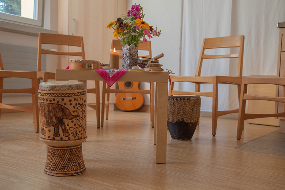 Das Bild zeigt einen Tisch mit einer Kerze und einem Blumenstrauss darauf. Rund um den Tisch stehen Stühle. Im Bild zu sehen ist zudem eine Gitarre und zwei Trommeln.