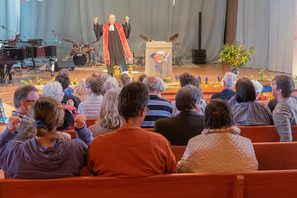 Das Bild zeigt einen Pfarrer auf einer Bühne. In den Bänken davor sitzen viele Menschen.