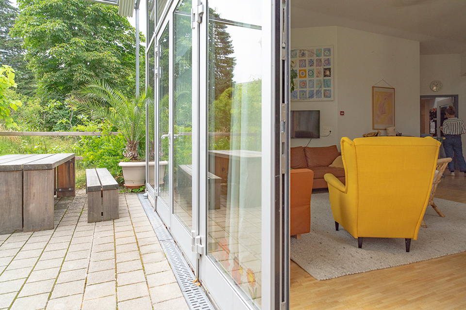 Das Bild zeigt in der einen Hälfte einen Gartensitzplatz mit einer Palme und in der anderen Hälfte ein Wohnzimmer mit einem markanten gelben Sessel.