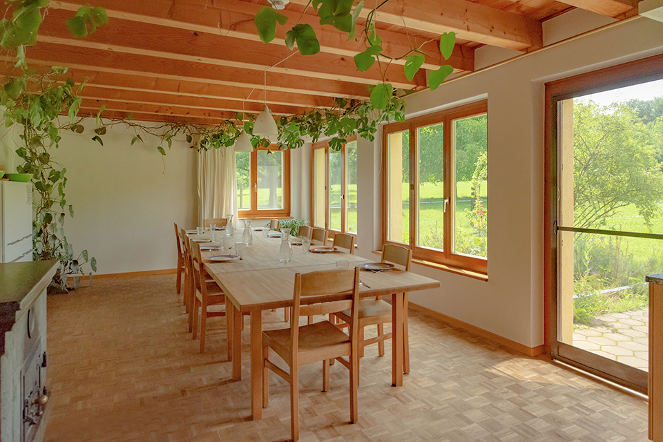 Das Bild zeigt einen grossen gedeckten Esstisch. Das Zimmer hat viele Fenster mit Sicht ins Grüne, einen Parkettboden und eine Decke mit Sichtbalken.
