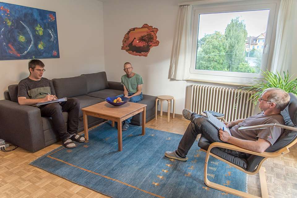 Das Bild zeigt drei Männer in einem Wohnzimmer die miteinander im Gespräch sind.