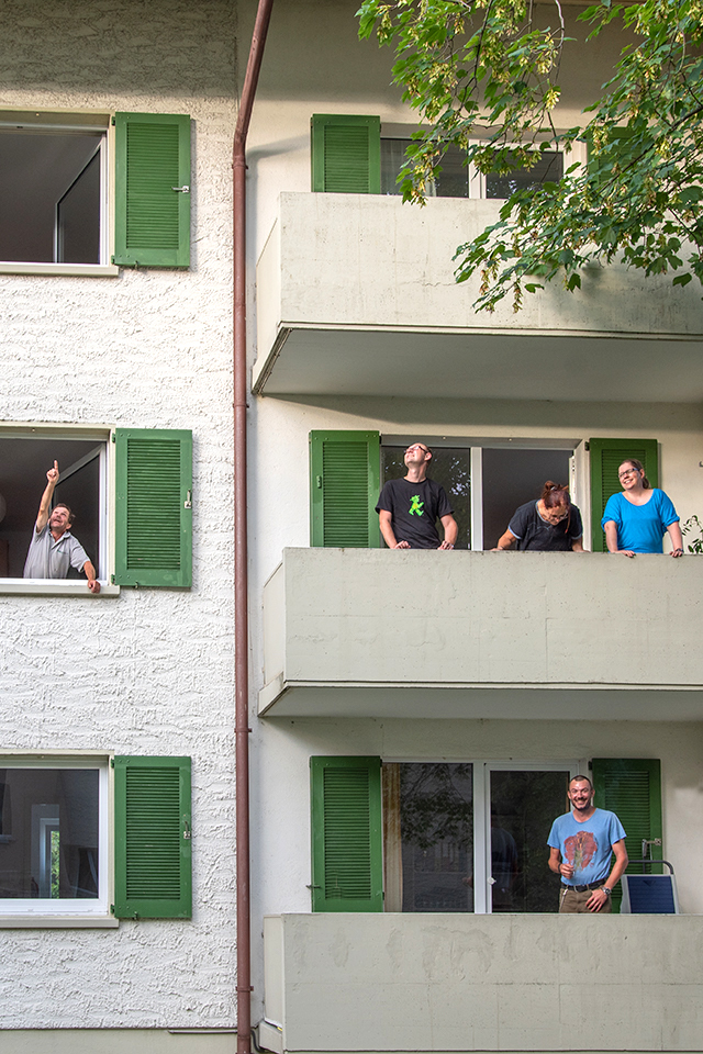 Das Bild zeigt einen Mann der aus dem Fenster eines Mehrfamilienhauses schaut und vier Personen auf einem Balkon stehend.