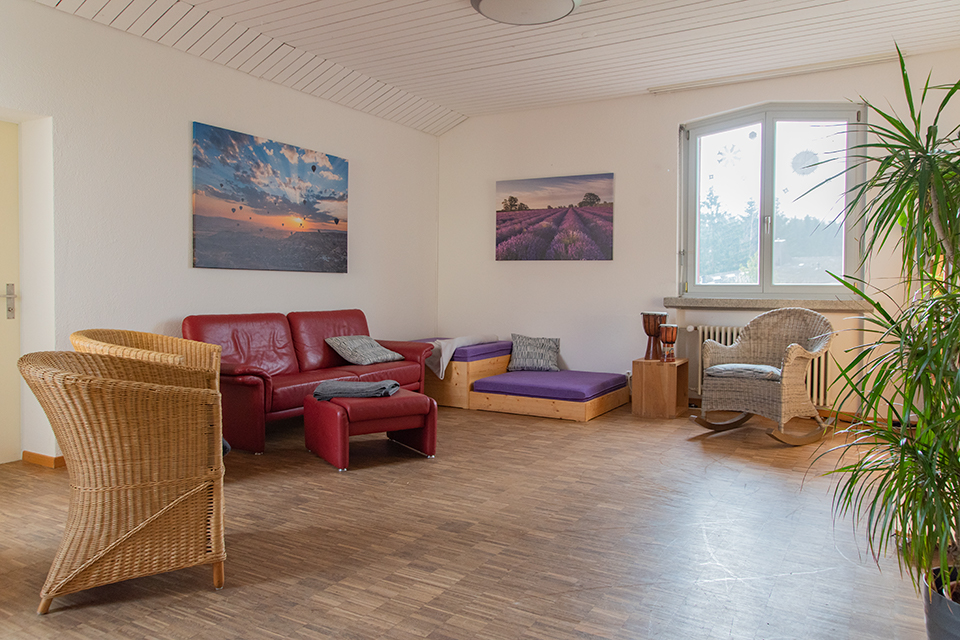 Das Bild zeigt ein Wohnzimmer mit rotem Sofa, Rattansesseln, Zimmerpflanzen und einem Podest zum draufliegen.