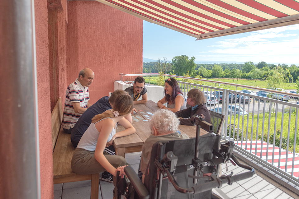Das Bild zeigt eine Gruppe Menschen auf dem Balkon am Esstisch sitzen beim Memoryspielen.