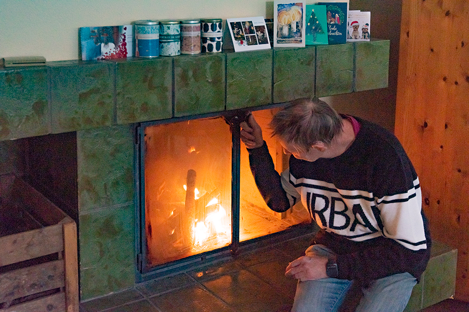 Das Bild zeigt einen Mann der im Wohnzimmer vor dem Kamin sitzt und das Feuer anschaut. Auf dem Kaminsims befinden sich viele Weihnachtskarten.