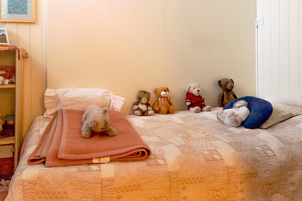 Das Bild zeigt ein Bett das mit einer hellen Tagesdecke zugedeckt ist. Auf dem Bett befinden sich viele Stofftiere und eine Wolldecke.