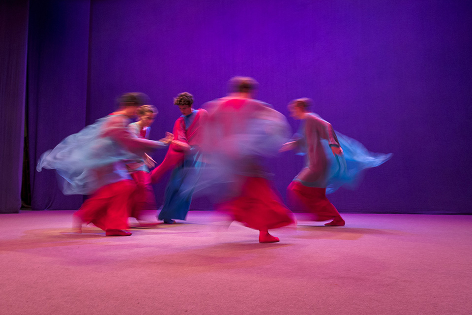 Das Bild zeigt fünf Tänzer in rotblauen Gewändern auf einer Bühne.