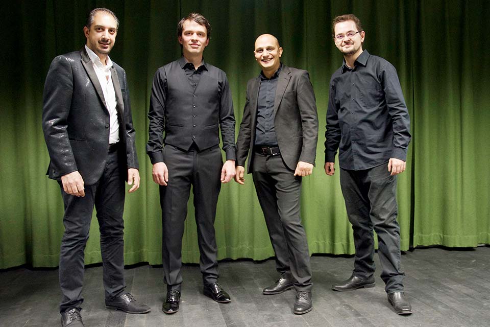 Das Bild zeigt vier Männer in schwarzen Anzügen vor einem grünen Vorhang stehen.