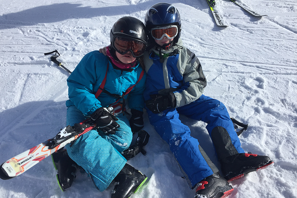 Das Bild zeigt zwei Jungen die in Skianzügen auf einer Skipiste sitzen.