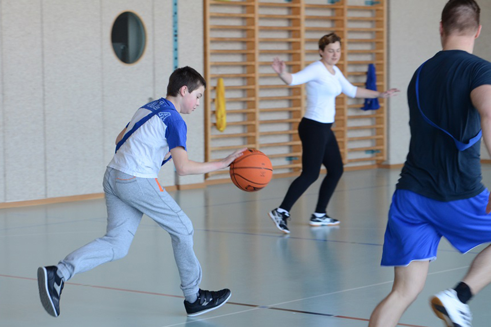 Das Bild zeigt einen Jungen der in einer Turnhalle zusammen mit anderen Kindern Basketballspielt.