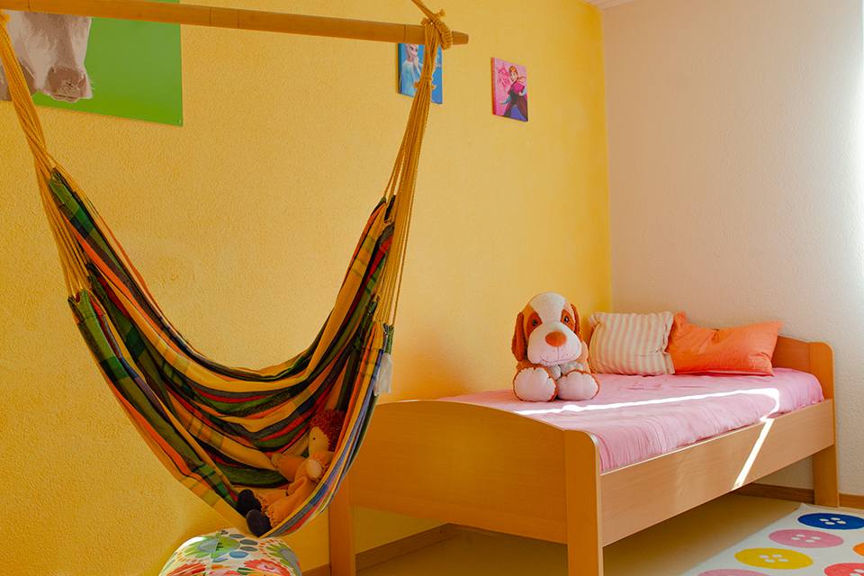 Das Bild zeigt ein Kinderzimmer mit gelben Wänden in dem ein Bett steht auf dem ein Plüschhund sitzt. In dem Zimmer befindet sich zudem eine Hängematte.