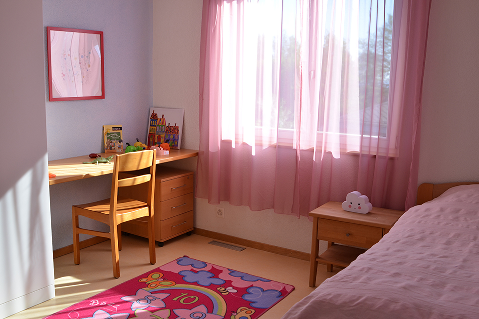 Das Bild zeigt ein Kinderzimmer mit rosaroten Vorhängen und ebensolcher Bettwäsche. In dem Zimmer befindet sich nebst dem Bett auch ein Schreibtisch.