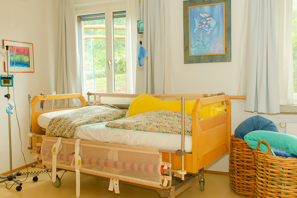 Das Bild zeigt ein Pflegebett gelbgrüner Bettwäsche. An der Wand hängt ein Bild mit einem Regenbogenfisch.