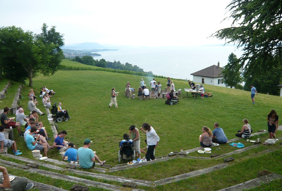Das Bild zeigt viele Menschen die auf einer Wiese essen. Im Hintergrund ist der Neuenburgersee zu sehen.