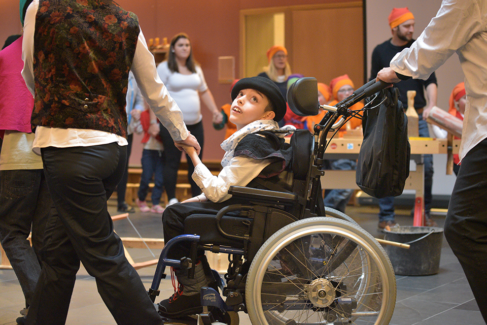 Das Bild zeigt einen Jungen im Rollstuhl. Zwei erwachsene Personen halten ihn an den Händen und Bilden so zusammen mit vielen weiteren Personen einen grossen Kreis.