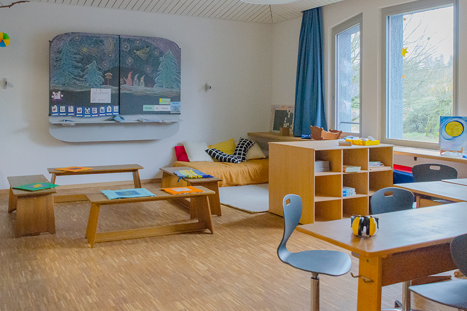 Das Bild zeigt ein Unterrichtszimmer mit einer Wandtafel, einem Sofa, einem Regal, mehreren Sitzbänken und einem Pult.