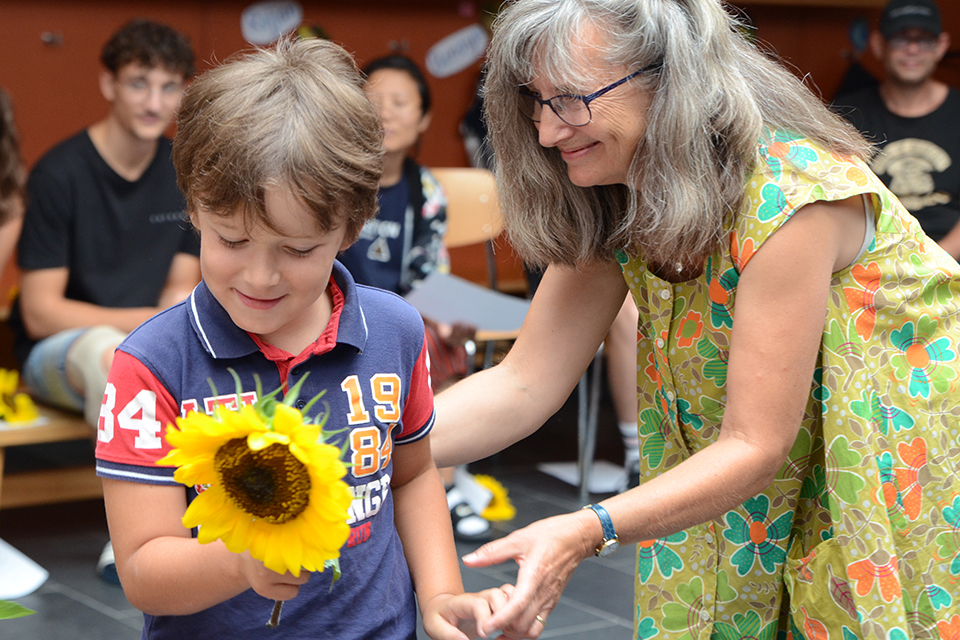 Das Bild zeigt einen Jungen mit einer Sonnenblume in der Hand. Neben ihm steht eine Frau die ihn anlächelt.