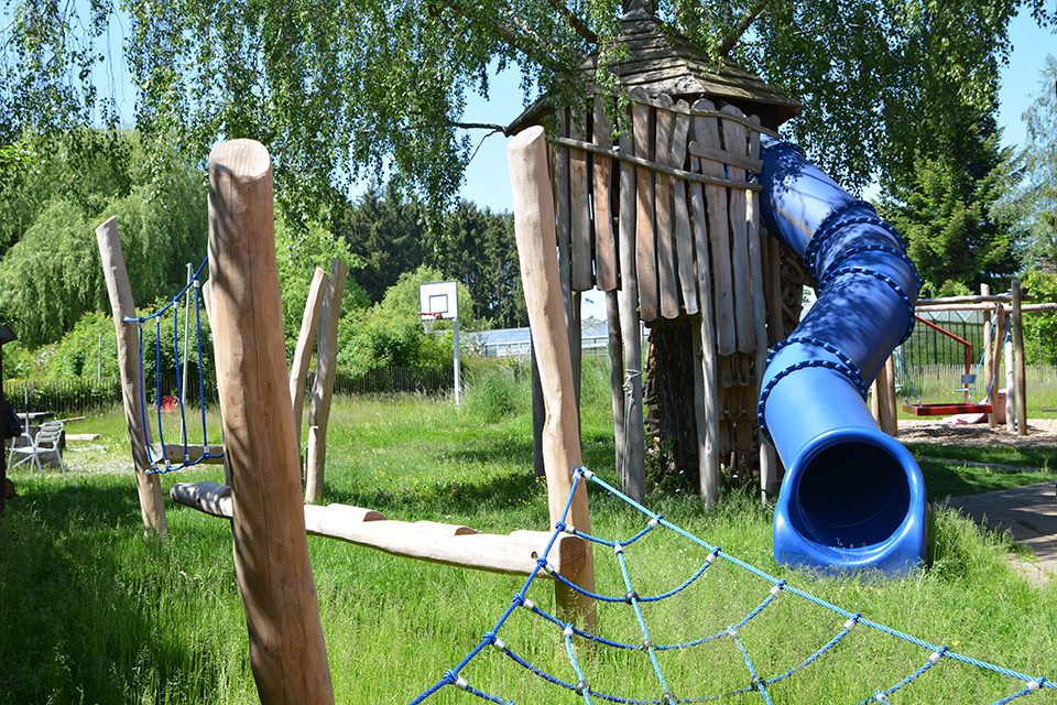 Das Bild zeigt ein Klettergerüst aus Baumstämmen und Seilen, ein Holzspielhaus mit blauer Rutsche und ein Basketballkorb.
