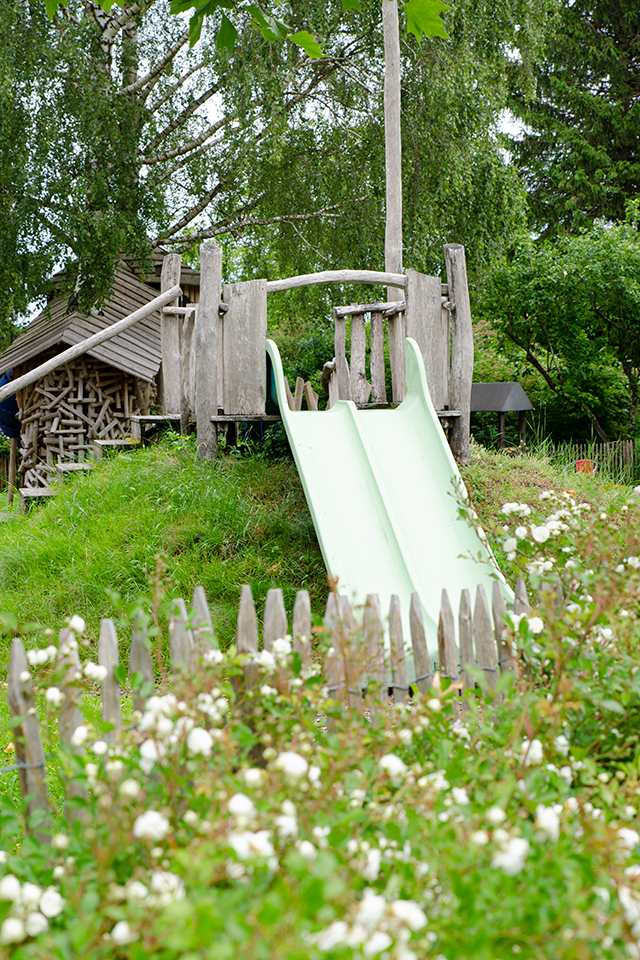 Das Bild zeigt ein grüner Wiesenhang auf dem eine Holzplattform steht und von der eine grüne Doppelrutschbahn wegführt.