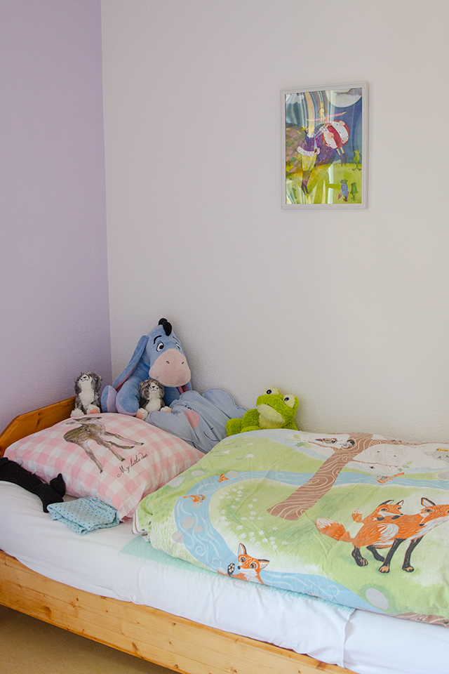 Das Bild zeigt ein Kinderzimmer mit weissen und violetten Wänden. In dem Zimmer steht ein Bett mit bunter Bettwäsche und Stofftieren darauf.