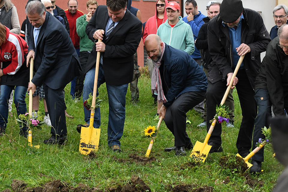 Das Bild zeigt fünf Männer die mit einer gelben Schaufel eine Wiese umgraben. Im Hintergrund stehen viele Schaulustige.