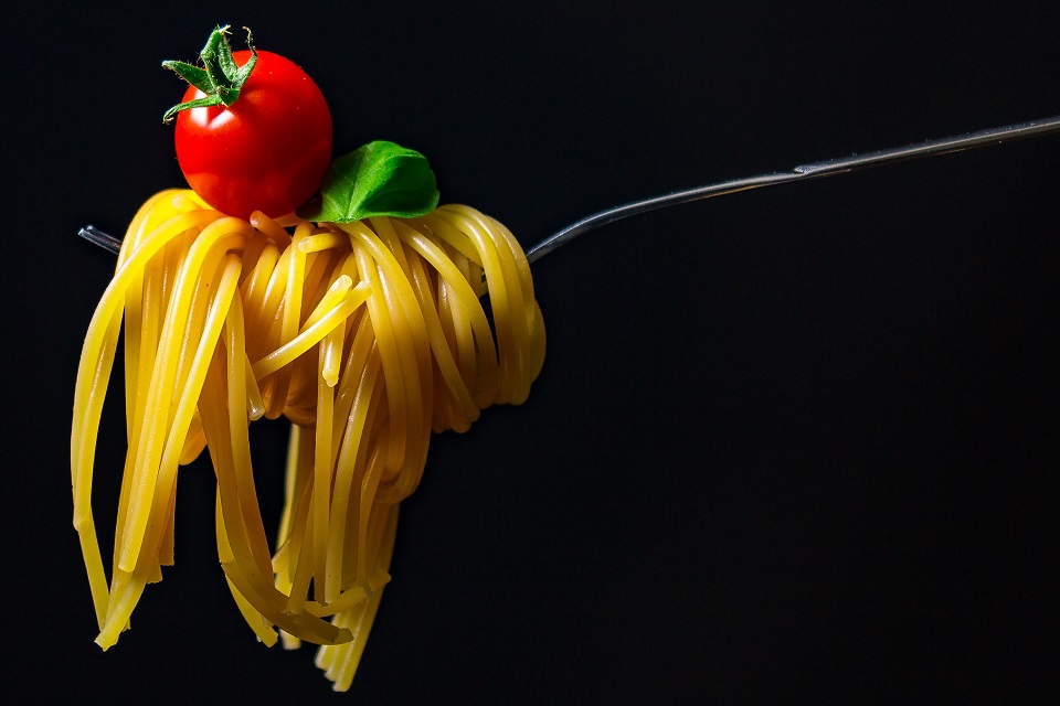 Das Bild zeigt eine Gabel mit Spaghetti, Cherrytomate und einen Blatt Basilikum.