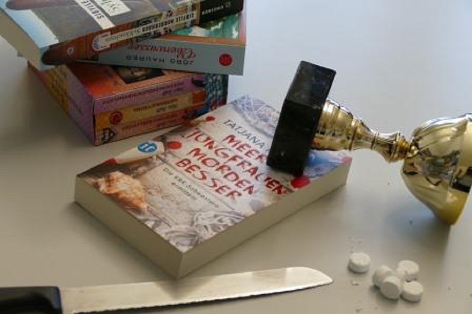 Das Bild zeigt mehrere Kriminalromane aufeinandergestapelt. Daneben liegen Tabletten, ein Küchenmesser und ein Pokal.