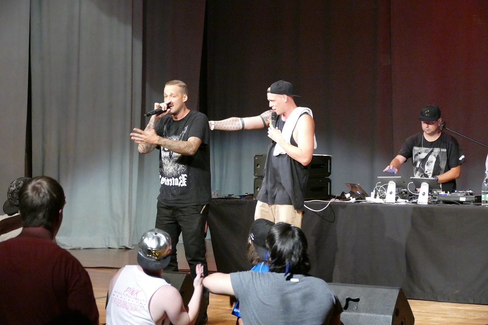 Das Bild zeigt drei Männer auf einer Bühne. Ein DJ und zwei Sänger.