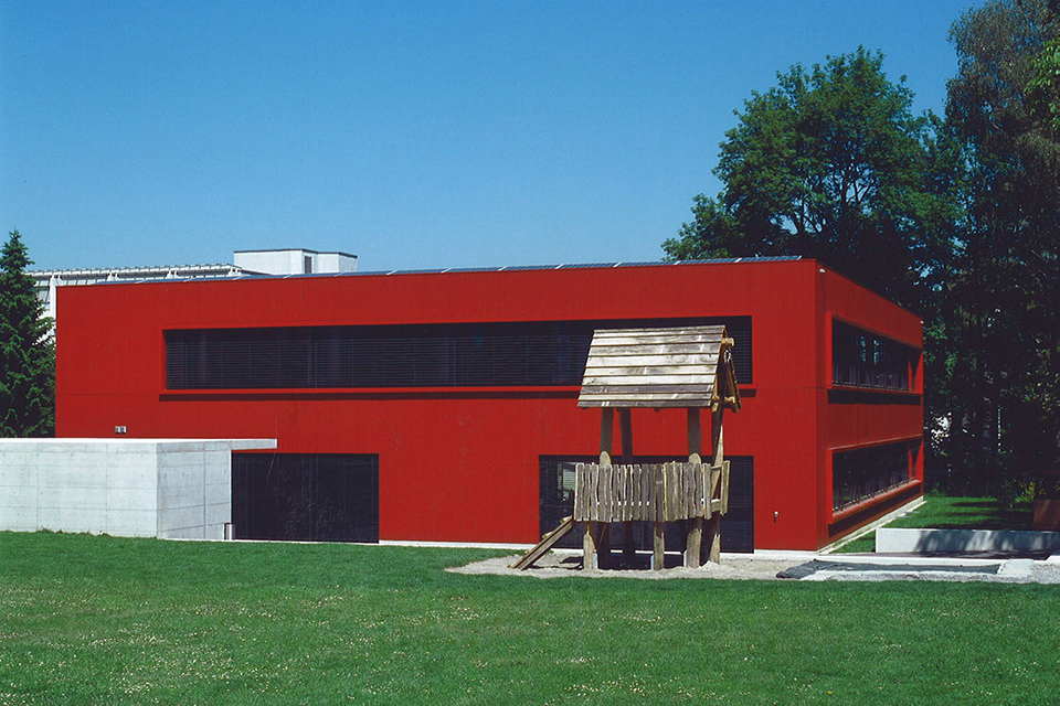 Das Bild zeigt eine rotes Gebäude mit Fenster die von schwarzen Rolländen bedeckt sind. Vor dem Gebäude steht ein Holzspielhaus.