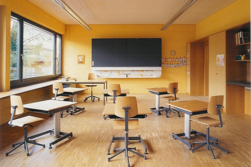 Das Bild zeigt einen grossen Raum mit Holzboden und gelben Wänden. In dem Raum befinden sich sechs Schulbänke, ein Lehrerpult und eine Wandtafel.