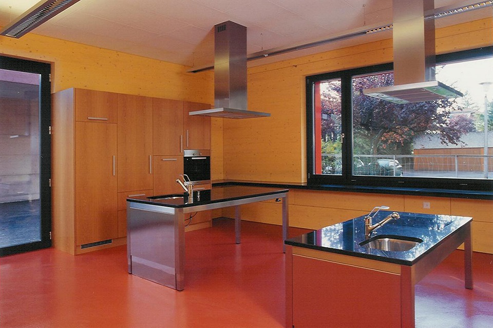 Das Bild zeigt eine Schulküche mit rotem Boden und gelben Wänden. An der Decke sind Dampfabzüge angebracht. In der Mitte des Raumes steht zwei Kochinseln.