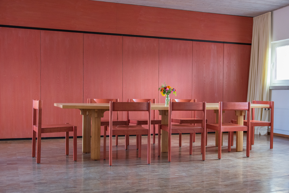 Das Bild zeigt einen grossen Holztisch mit 8 roten Stühlen vor einer roten Holzwand.