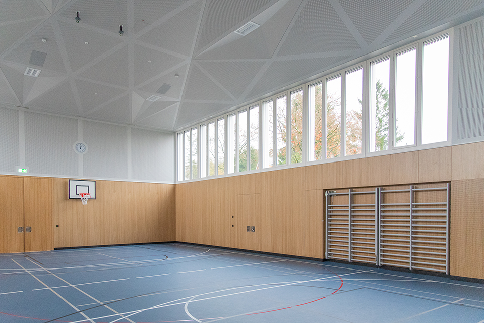 Das Bild zeigt eine Turnhalle mit blauem Boden, einer Holzwand und hochangesetzten Fenstern. An den Wänden befinden sich vier Sprossenwände und ein Basektballkorb.