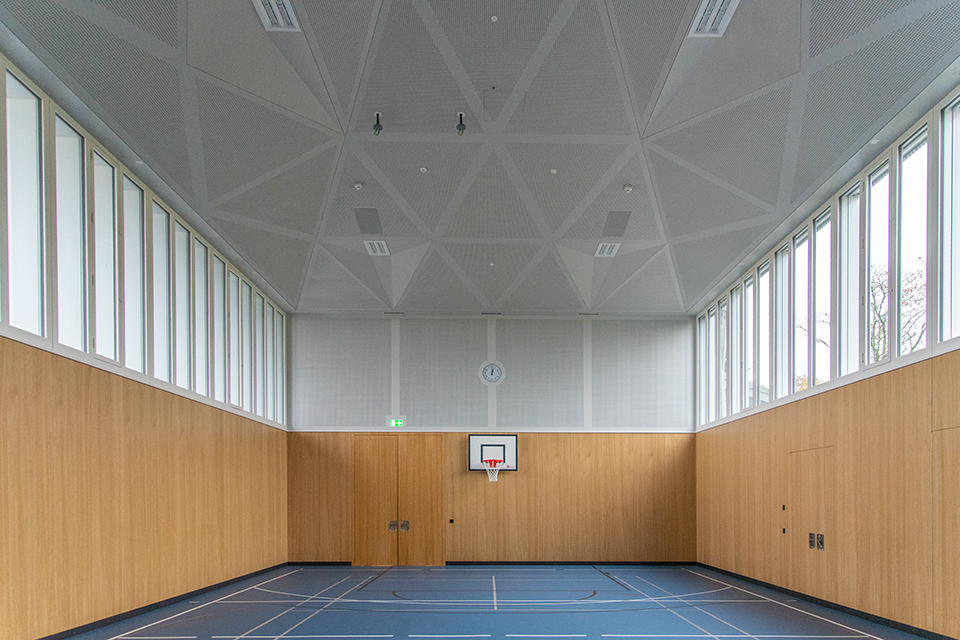Das Bild zeigt eine Turnhalle mit blauem Boden, einer Holzwand und hochangesetzten Fenstern. An der Wand befindet sich ein Basektballkorb.